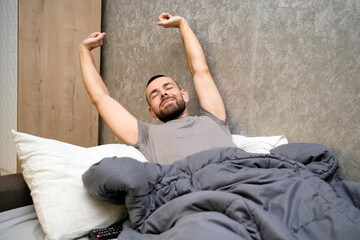 A man is waking up in bed, stretching his arms above his head. He has a relaxed expression on his face. The blanket is draped over him as he enjoys a moment of rest after being unwell