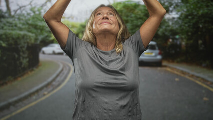 Senior blonde woman raising bare arms overhead in a gray t shirt on a residential street, smiling with parked car, hedge and trees visible; joy resilience.