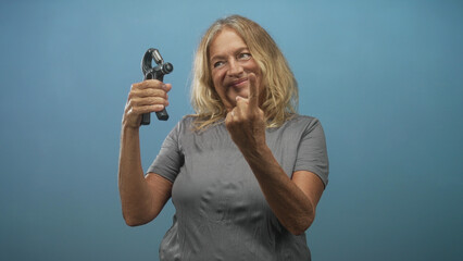 Woman blonde wearing gray tee holding hand exerciser and showing middle finger to camera in studio; playful defiance strength.