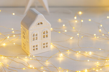 Soft Christmas scene with white ceramic house, glowing warm fairy lights and neutral winter decor....