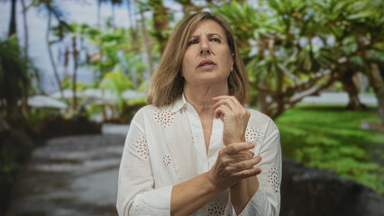 Fototapeta premium Woman in white blouse touches wrist while squinting and examining hand amid green foliage in forest; concern.
