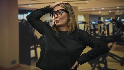 Fototapeta premium Woman with hand on head and mouth open wearing glasses in gym with cardio machines and bench visible; surprise playful determination.
