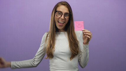 Woman wearing glasses and white top holding a pink sticky note and pointing finger to it in studio with purple backdrop; happiness optimism.