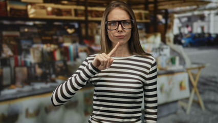 Woman pointing index finger toward camera in a street market stall, striped shirt and glasses, hand raised in a clear warning gesture; firm disapproval.