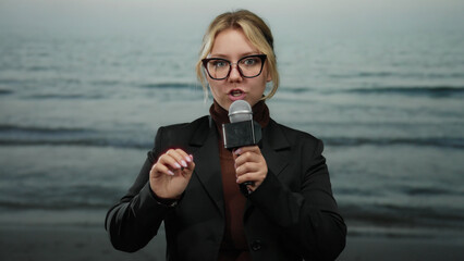 Young woman reporter with microphone speaking on beach seaside with sea background under clear sky...