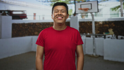 Man smiling and standing beside a basketball hoop on a street court wearing red tshirt and looking at camera; youthful joy.