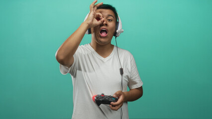 Hispanic man holding game controller and wearing headset while making ok sign by eye in turquoise studio; playful focus.
