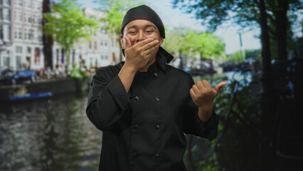 Man chef in black uniform with hand over mouth pointing thumb to street canal in amsterdam near...