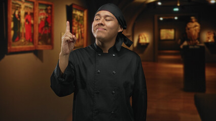 Young hispanic man chef in black uniform and headscarf points finger upward in an art gallery building, smiling with raised right hand; confidence art appreciation.