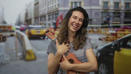 Young hispanic woman smiling while hugging a ukulele with her hands on a busy city street lined with taxis and construction barriers; joy.
