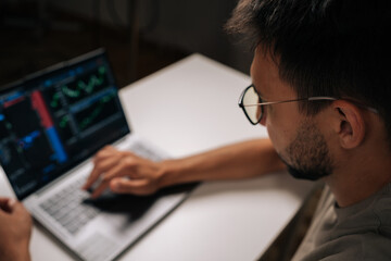 View from shoulder of trader in glasses typing on laptop keyboard, engaging in online trading and financial analysis, viewing real-time charts and data for informed investment decisions at home.