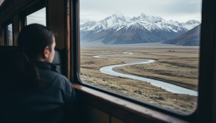 Pensive woman looking out a train window at a majestic snow-capped mountain range and a winding river in a vast, scenic valley during her journey