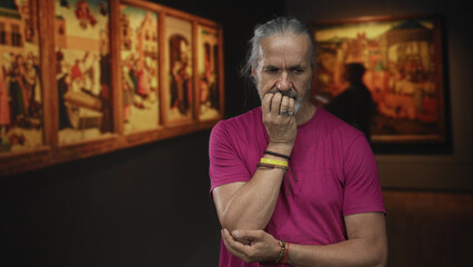 Man biting nails with hand to mouth, long grey hair and rings in an art gallery building beside...