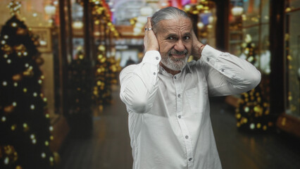 Fototapeta premium Man with gray hair and beard covers ears in decorated shopping building corridor holding hands to head in pain; holiday discomfort.