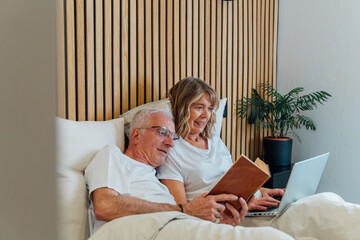 Senior couple relaxing comfortably in bed together, man reading a book while woman uses a laptop, both engaging in leisure activities and unwinding at home