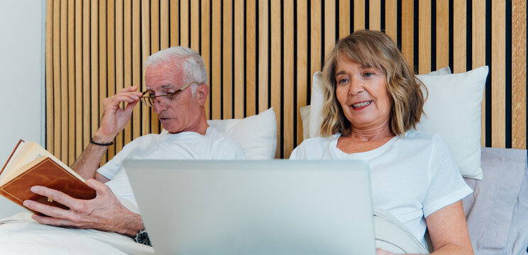 Senior couple relaxing in bed, man reading a book, woman using a laptop, enjoying leisure time and modern technology together at home