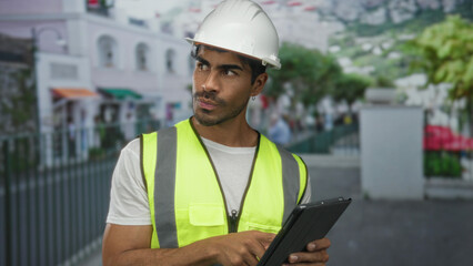 Hispanic man taps tablet on street as engineer wearing yellow reflective vest and white hardhat; determination.
