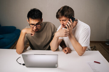 Portrait of two young entrepreneurs in home office collaborate on startup project, one speaks on phone while other concentrates working on laptop, planning and problem-solving.