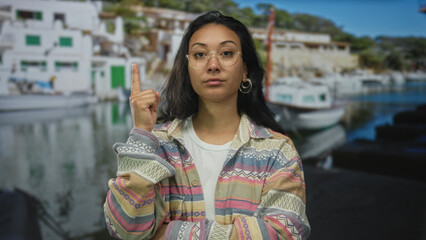Woman with raised index finger pointing up beside boats at port marina wearing glasses and patterned jacket; assertion confidence.