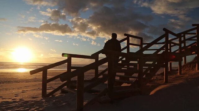 Lone traveler ascends wooden stairs on a Spanish beach, Cuesta Maneli, Huelva at sunset sunrise, silhouetted against the dramatic golden sky. Travel, peace, adventure.