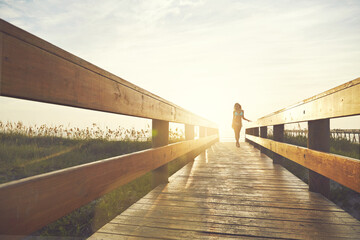 Obraz premium Woman, boardwalk and walking at beach with sunshine, below space and vacation for summer travel. Adventure, portrait or person in bikini for coastal holiday, sunrise sky and relax by wood pathway