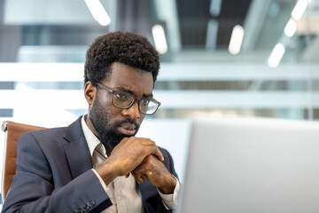 Businessman wearing eyeglasses and a suit, sitting in an office chair, deeply concentrating and thinking while working on a laptop in a modern corporate setting
