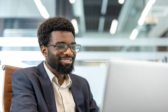African american businessman smiling at laptop in a modern, sunlit office, focused on digital work confident, professional manager portraying productivity, tech savvy success