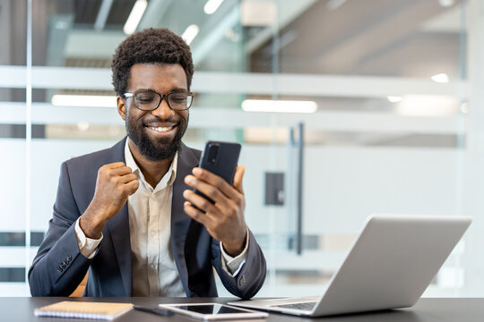 African american businessman receiving good news on mobile phone, showing happiness and celebrating victory with a fist pump while working at a modern office desk