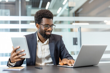 African american businessman multitasking while using a laptop and digital tablet at his office desk, reflecting modern business communication and productivity