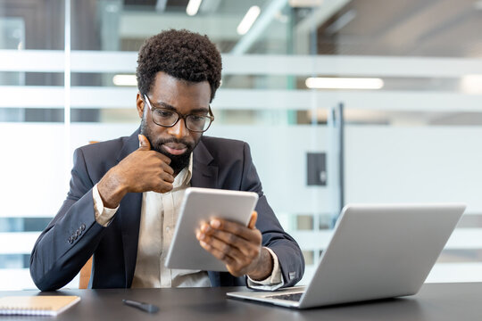African american businessman sitting at desk, concentrating on digital tablet, making important decisions and utilizing modern technology in a corporate office environment - Powered by Adobe