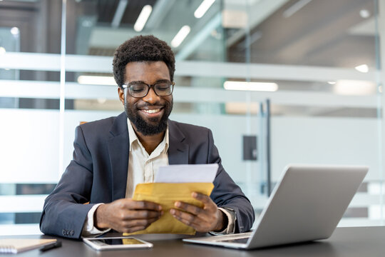 Smiling young businessman reading official documents from an envelope in an office, celebrating success and happy news after hard work and dedication