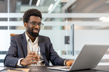 Fototapeta premium Businessman in smart-casual suit smiles while working on a laptop at a modern corporate desk, holding a glass of water, confident and focused during remote work
