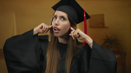 Woman wearing black graduation gown and mortarboard plugs ears with fingers in building; annoyance.