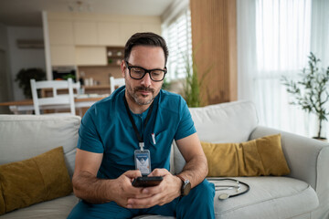 Male doctor in scrubs using smart phone on living room sofa