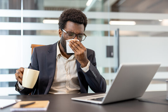 African american businessman at his desk feeling unwell during a cold or flu, blowing his nose with a tissue while holding a warm mug and working on his laptop - Powered by Adobe