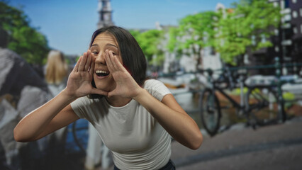 Fototapeta premium Young hispanic woman with hands cupped around mouth shouting on a street in amsterdam; excitement announcement.