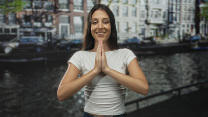 Young hispanic woman pressing hands together in prayer on an amsterdam canal street with buildings behind her; mindfulness gratitude serenity.