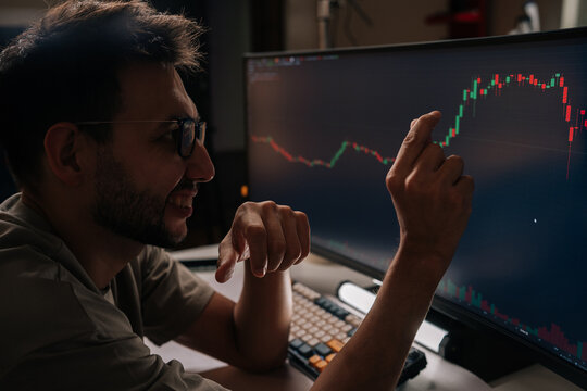 Positive young man smiling and pointing at fluctuating financial graph on desktop screen, actively managing cryptocurrency investments and tracking market trends for profitable trading decisions.