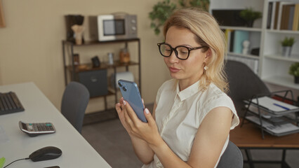 Woman wearing glasses and white blouse holds smartphone and scrolls screen inside modern building; productivity.