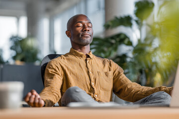 Black man doing a desk yoga twist in a modern office. Calm, inclusive wellness moment with natural light and greenery. Man performing subtle desk yoga twist, bright office, work-life balance wellness 