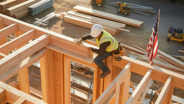 A photograph of a construction worker in a hard hat and high-visibility vest framing the wooden structure of a new house. The builder is positioned on scaffolding, carefully measuring and securing woo