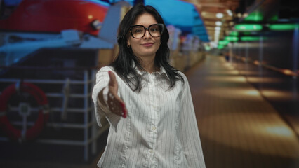 Fototapeta premium Woman wearing glasses and white shirt extending hand for handshake inside building; welcome connection.