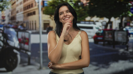 Young hispanic woman resting chin on hand in bright sunlit early morning urban street; serenity reflection.