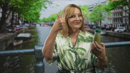Woman inspects and holds a green juice bottle and gives a thumbs up on street near a canal and...