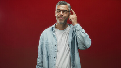 Man with glasses touches temple with index finger, smiling gently and wearing casual shirt against a red studio backdrop; thoughtful.