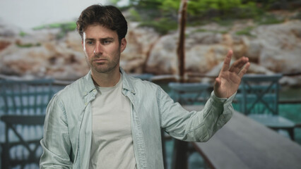 Young hispanic man points finger toward the sea from a building terrace at the coast, leaning over a wooden table and chairs; contemplation reflection calm.