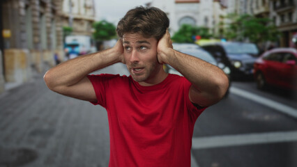 Young hispanic man in red t shirt covering ears with hands amid busy downtown street traffic and...