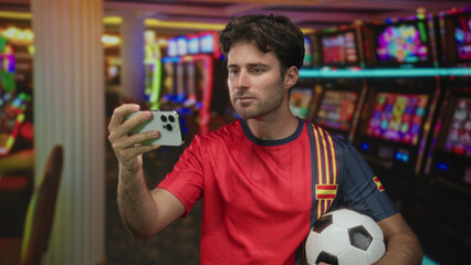 Man taking a selfie with smartphone while holding a soccer ball in a casino building; excitement fan pride.