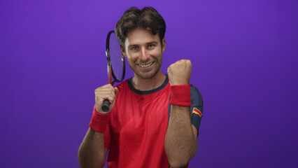 Young hispanic man holds tennis racket and makes a fist pump in studio with a purple backdrop while wearing red shirt and wristbands; victory momentum.