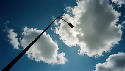 Tall dark street lamp silhouette stands against a dramatic deep blue sky with bright, sunlit clouds, captured from a low-angle perspective with the sun peeking through